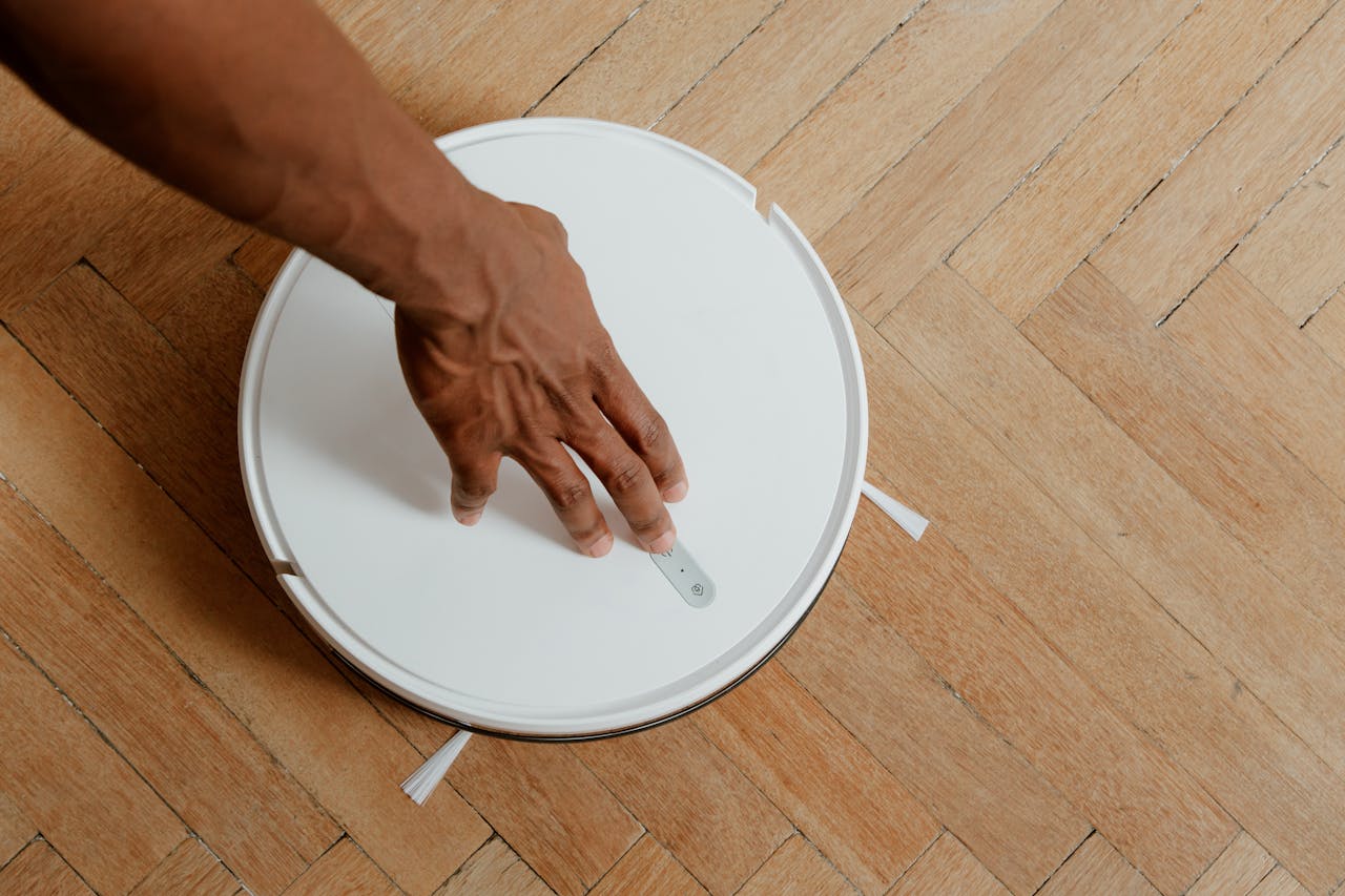 gallery-6 A close-up of a hand activating a robotic vacuum cleaner on a wooden floor.